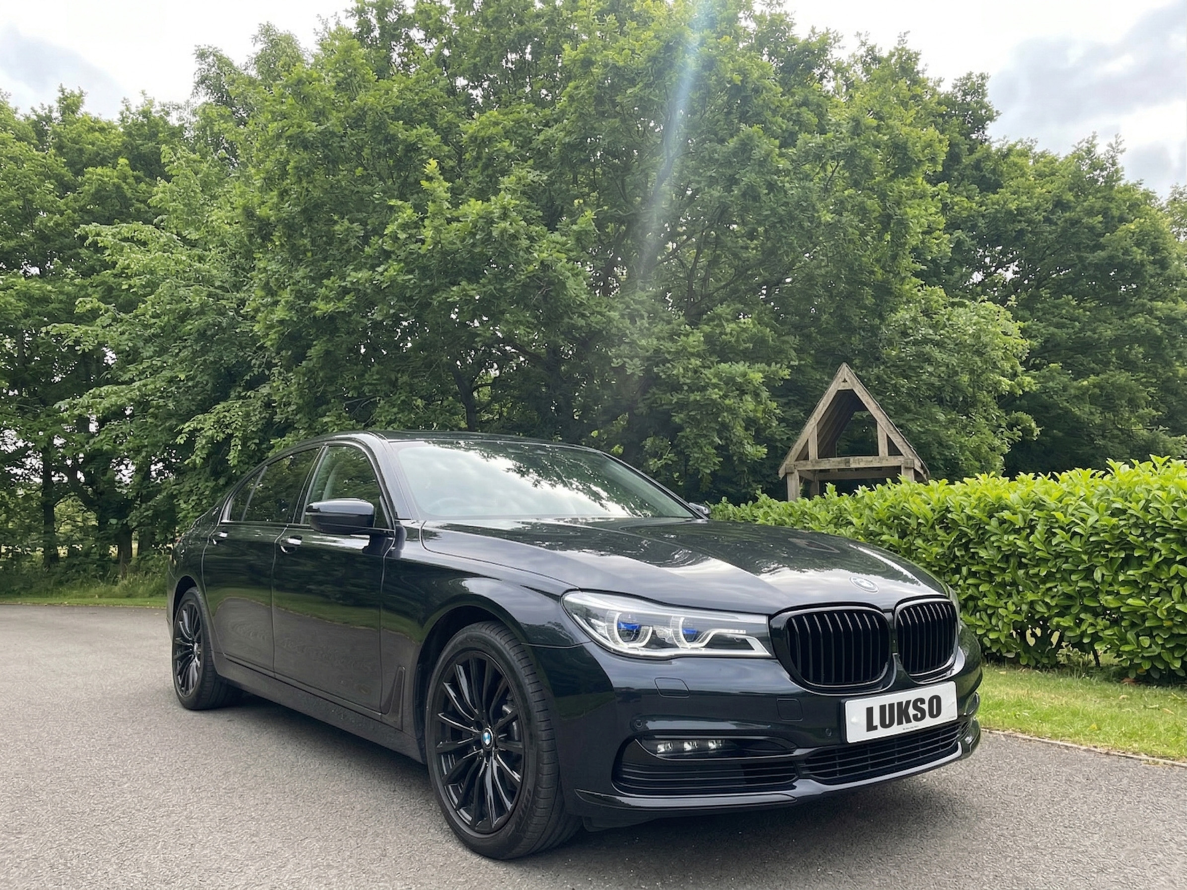 A black BMW saloon from our bespoke chauffeur service is parked on a tarmacked road near green trees and bushes, with a wooden structure in the background.