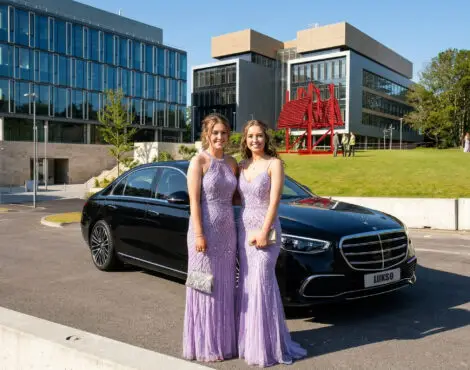 Two women in matching lavender dresses stand in front of a black prom car hire vehicle, with modern office buildings and a group of people in the background on a sunny day.