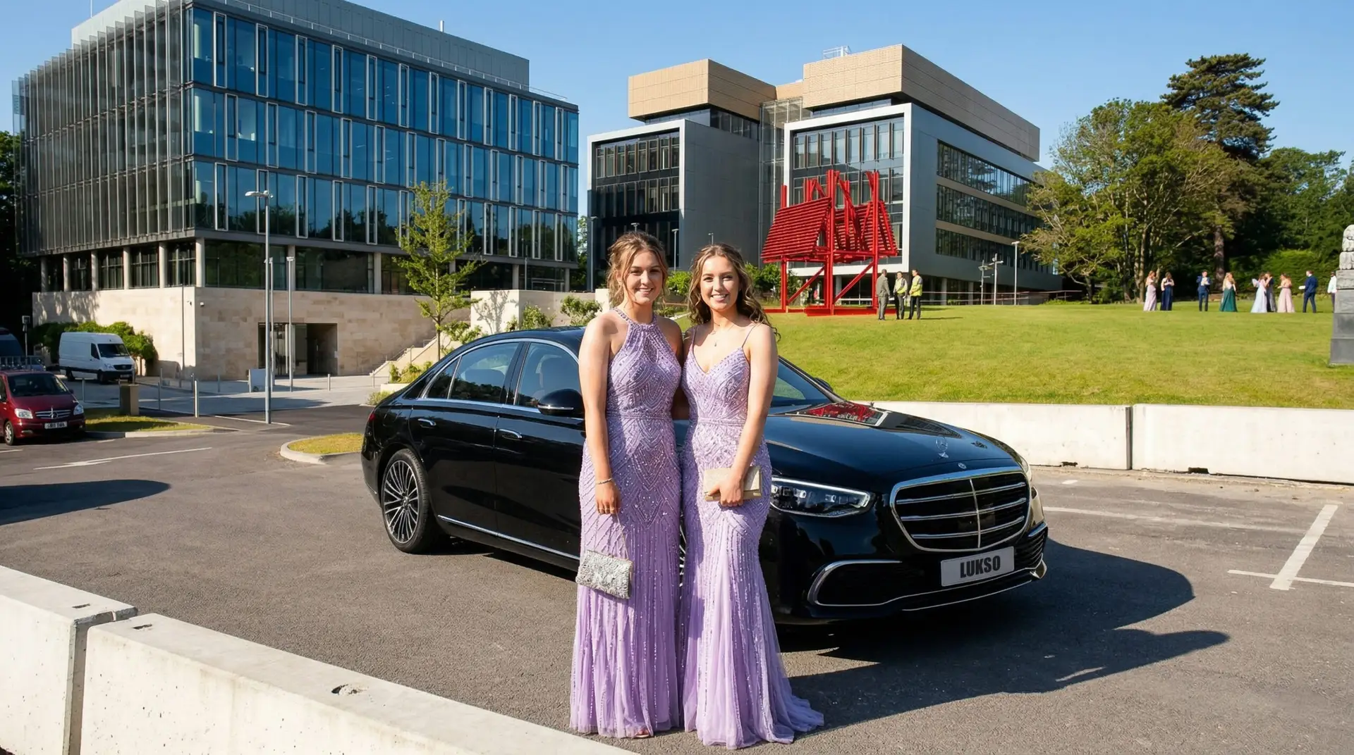 Two women in matching lavender dresses stand in front of a black prom car hire vehicle, with modern office buildings and a group of people in the background on a sunny day.
