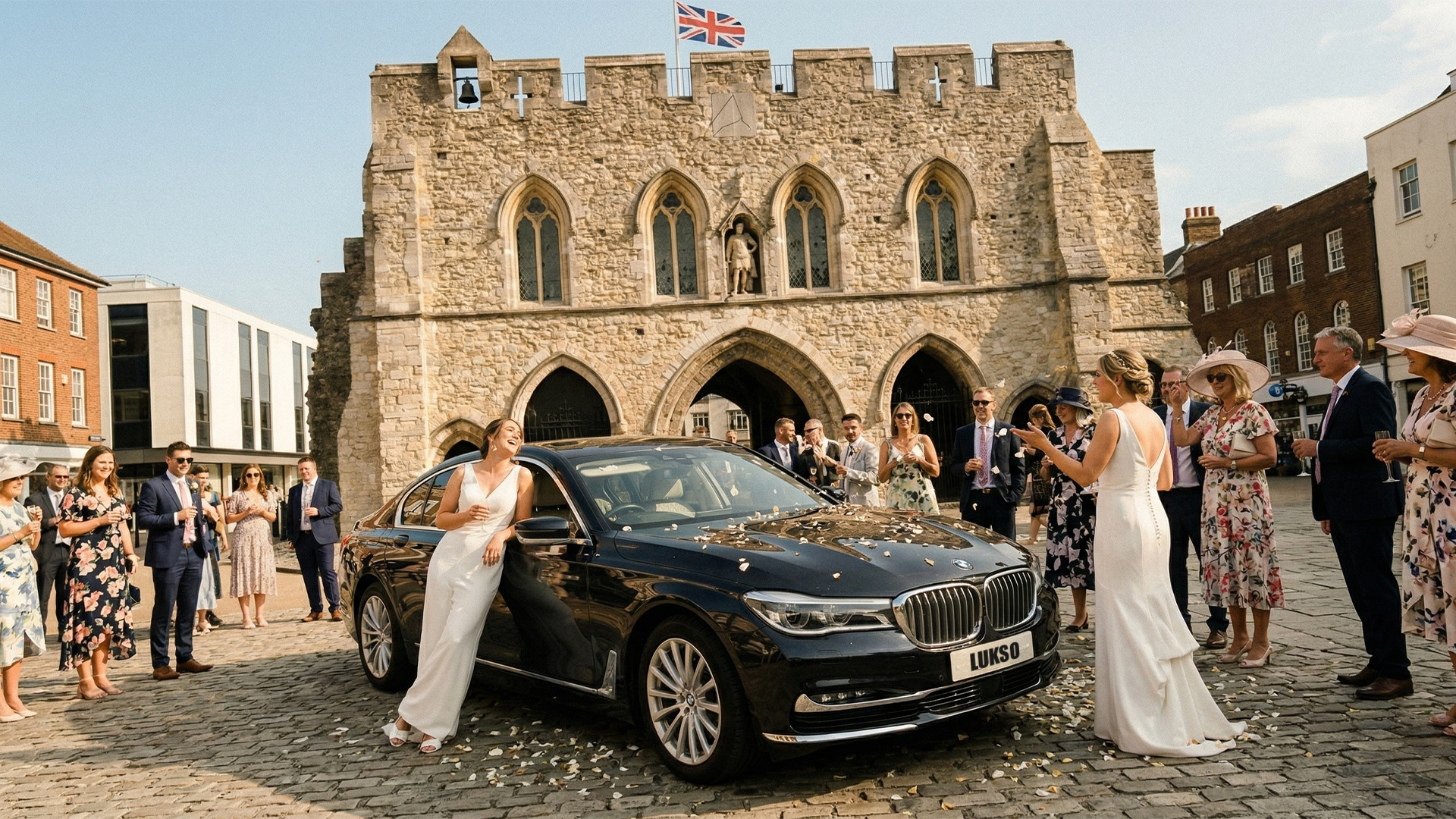 A bride in a white dress stands by a black BMW wedding car hire in front of a historic stone building, with guests surrounding her and flower petals scattered on the ground.