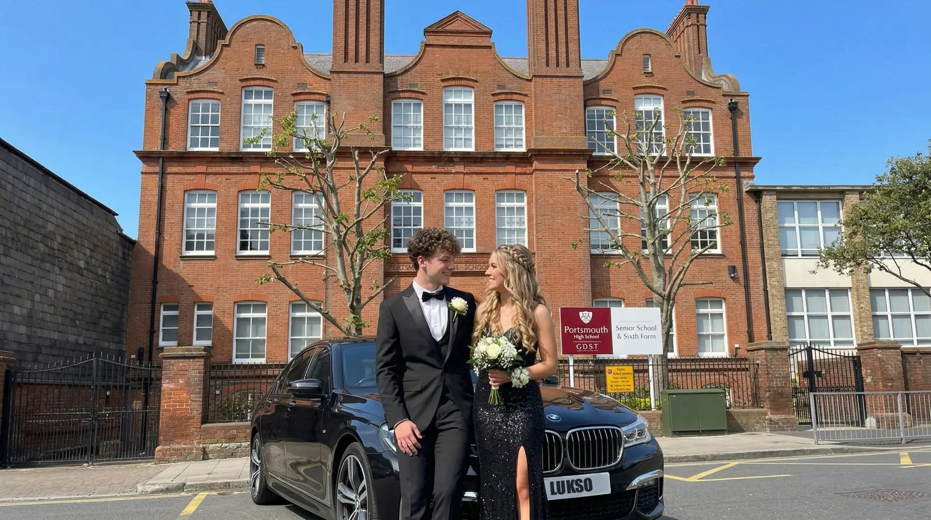 Two young adults dressed formally stand in front of a black car, holding flowers, as if ready for airport transfers, with a brick school building and a Portsmouth High School sign in the background.