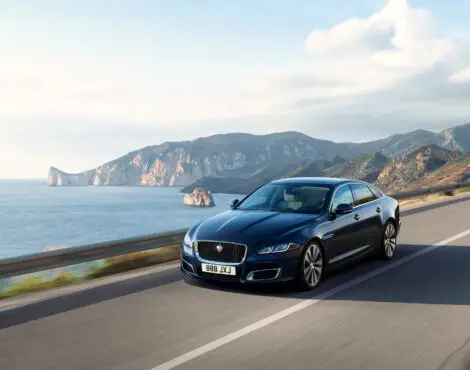 A black luxury saloon driven by a professional chauffeur Portsmouth glides along a coastal road, with cliffs, mountains, and the sea visible in the background under a partly cloudy sky.
