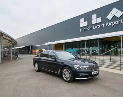 A black BMW 5 Series saloon is parked in front of the entrance to London Luton Airport on a cloudy day.