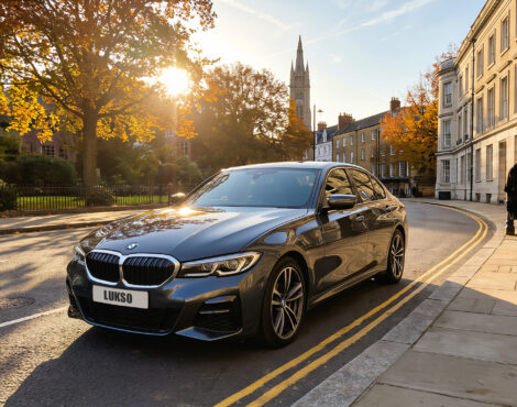 A grey BMW 5 Series saloon with the number plate LUKSO is parked on a sunlit street lined with historic buildings and autumn trees, as two people walk on the pavement.