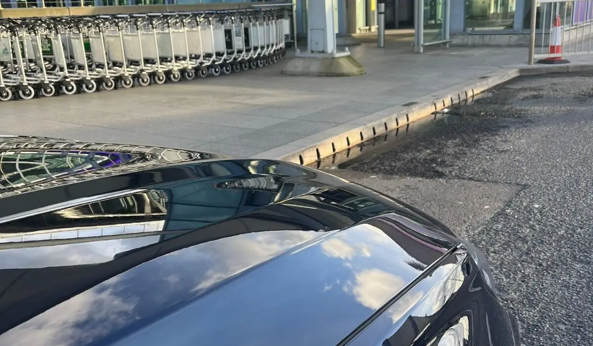 A close-up of a parked car’s bonnet reflecting the sky, with an airport entrance, luggage trolleys, and glass building in the background—perfect for a professional chauffeur Portsmouth arrival.