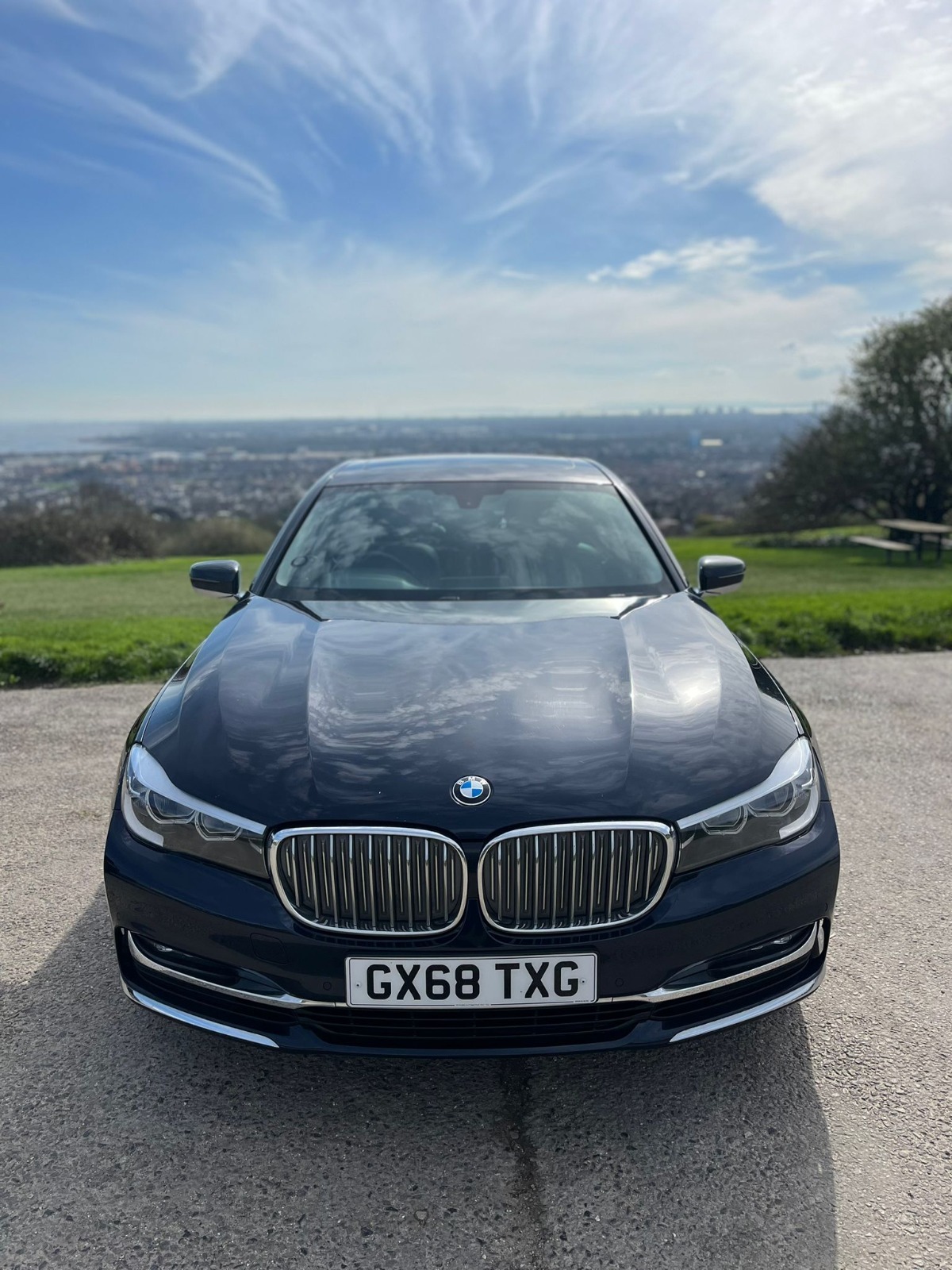 A dark blue BMW sedan with license plate GX68 TXG is parked on a hilltop, with a cityscape and cloudy sky visible in the background.