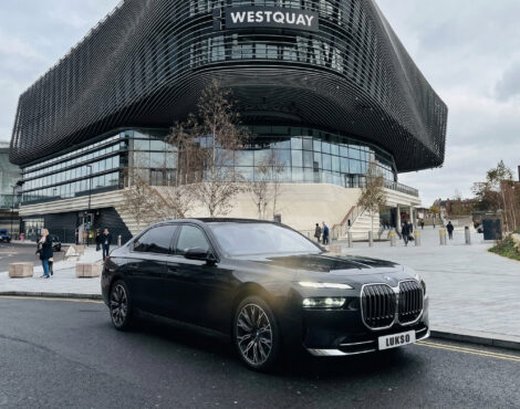 A black BMW 7 Series (LWB) saloon is parked on a street in front of the modern Westquay shopping centre, with people walking nearby.