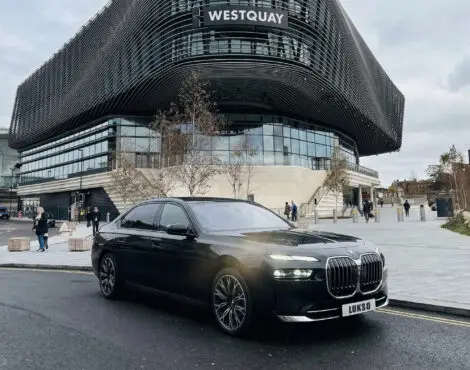 A black BMW 7 Series (LWB) saloon is parked on a street in front of the modern Westquay shopping centre, with people walking nearby.
