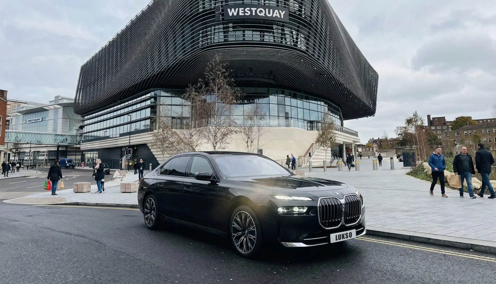 A black BMW 7 Series (LWB) saloon is parked on a street in front of the modern Westquay shopping centre, with people walking nearby.