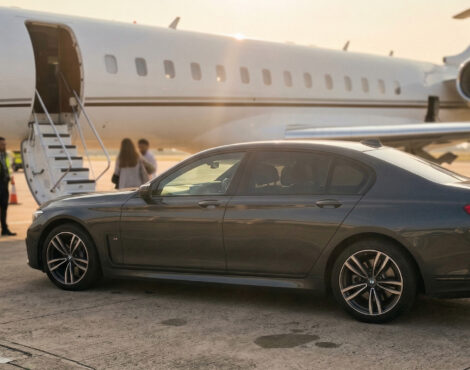 A black BMW 7 Series (LWB) saloon is parked on an airport apron in front of a private jet, with several people gathered near the jet’s steps in daylight.