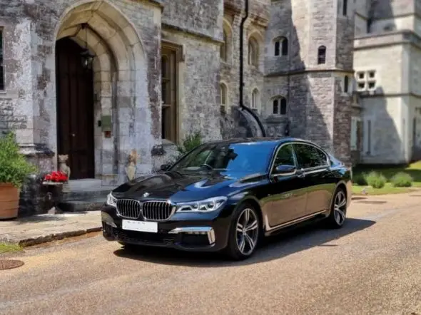 A black luxury saloon, ideal for a chauffeur Winchester experience, is parked on a paved driveway in front of a stone building with arched doorways and windows.