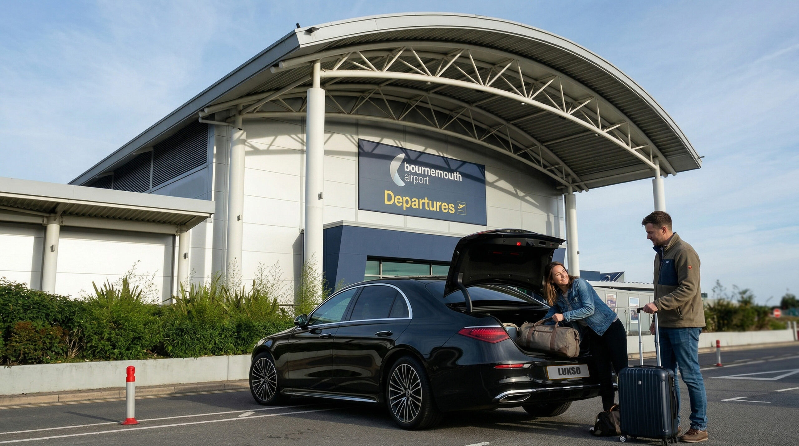 Two people unload luggage from a black Mercedes E-Class outside the departures area of Bournemouth Airport on a clear day.