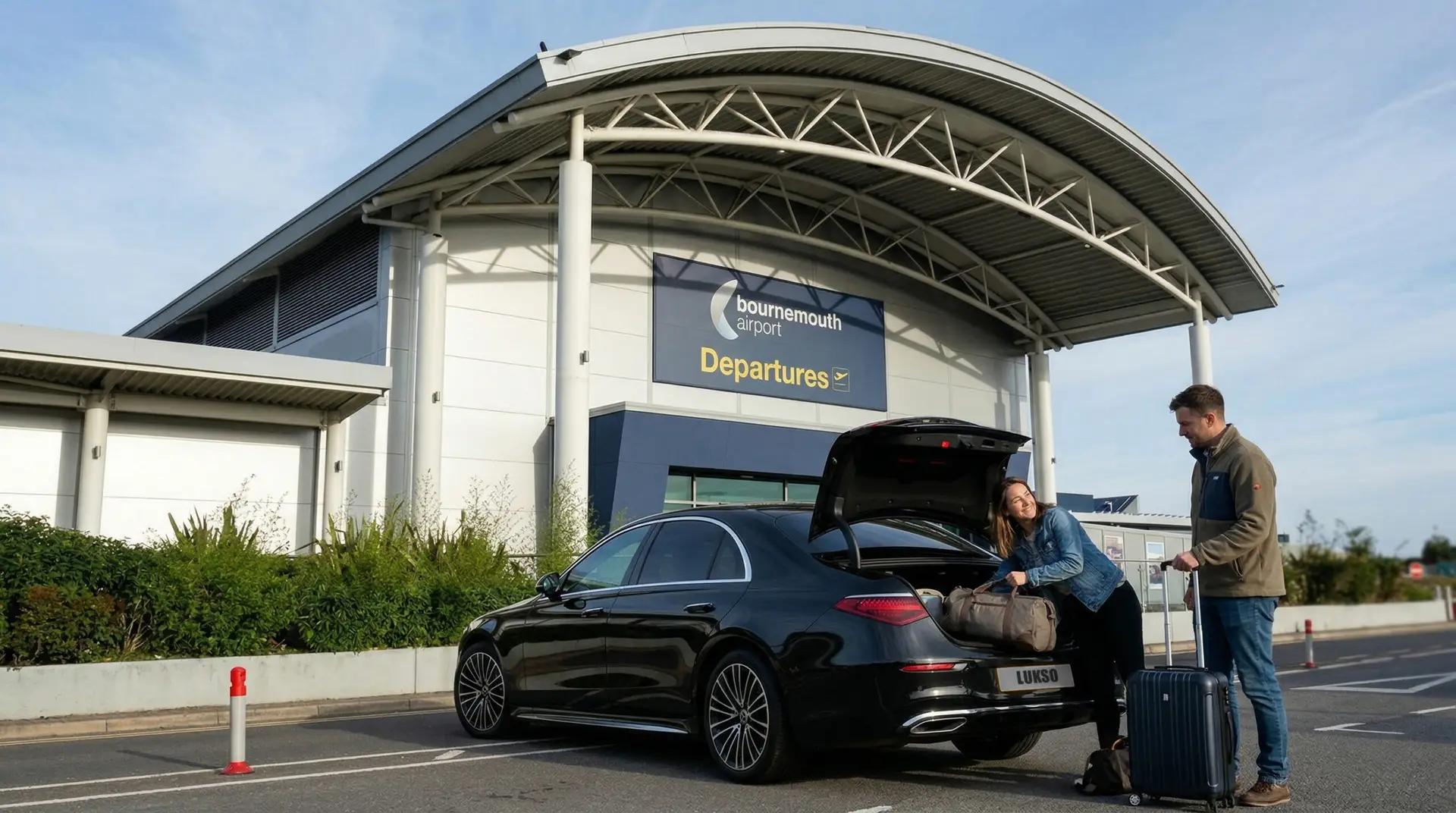 Two people unload luggage from a black Mercedes E-Class outside the departures area of Bournemouth Airport on a clear day.