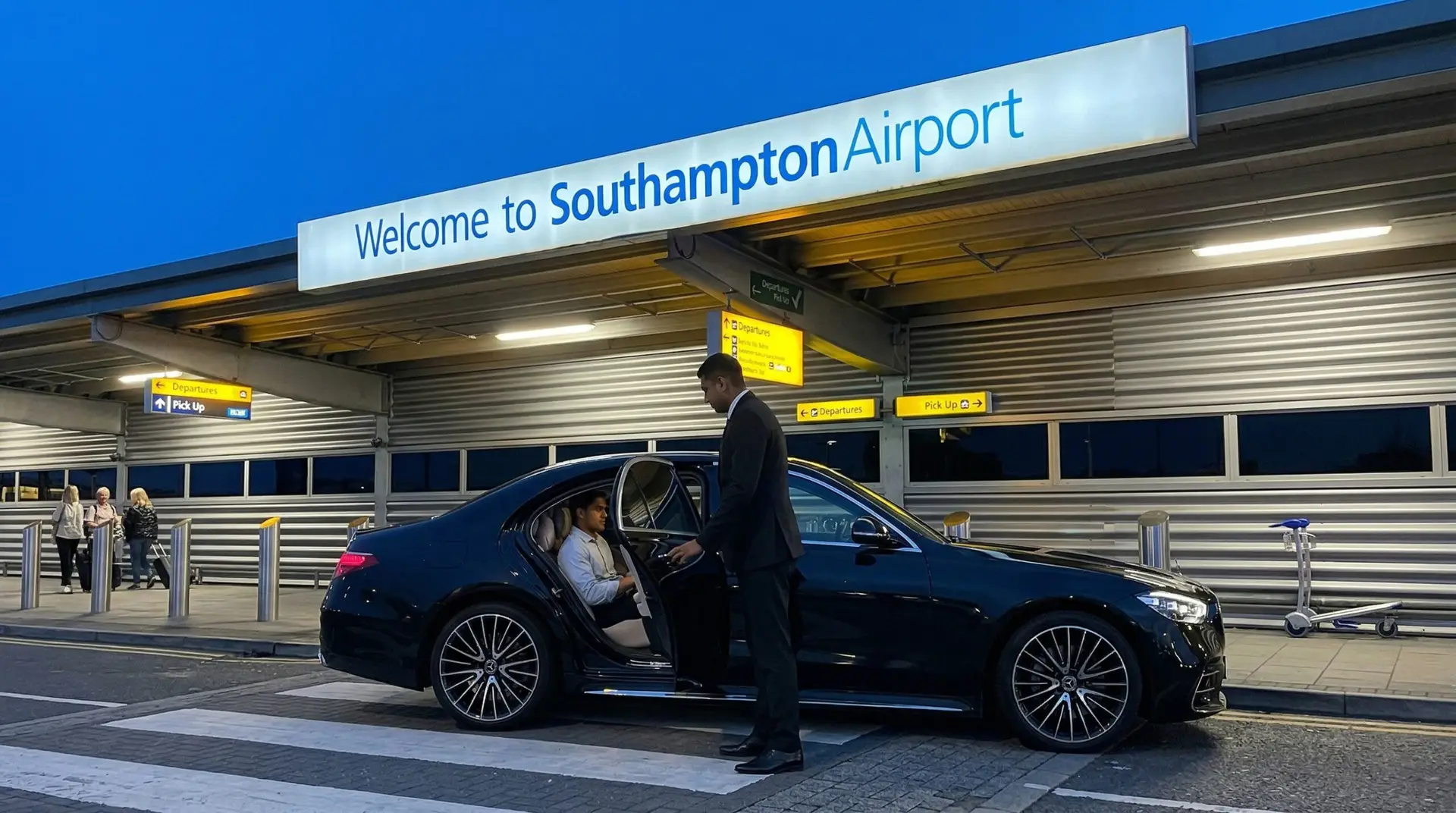 A chauffeur opens the door of a black Mercedes E-Class for a passenger outside the entrance to Southampton Airport at dusk.