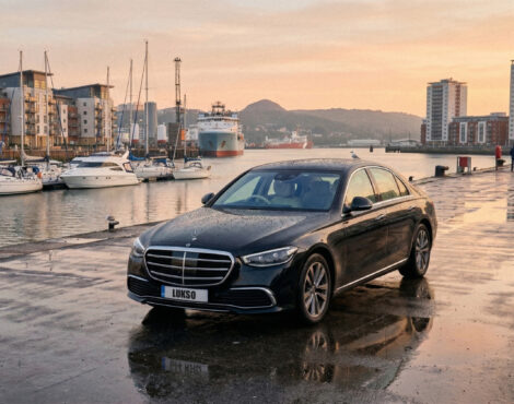 A black Mercedes S Class (LWB) saloon is parked on a wet quayside near boats and modern buildings at sunset.