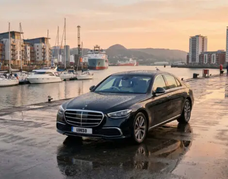 A black Mercedes S Class (LWB) saloon is parked on a wet quayside near boats and modern buildings at sunset.