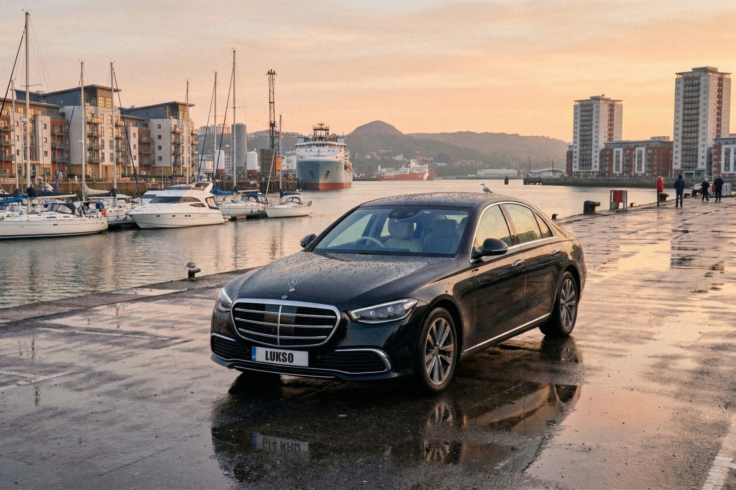 A black Mercedes S Class (LWB) saloon is parked on a wet quayside near boats and modern buildings at sunset.