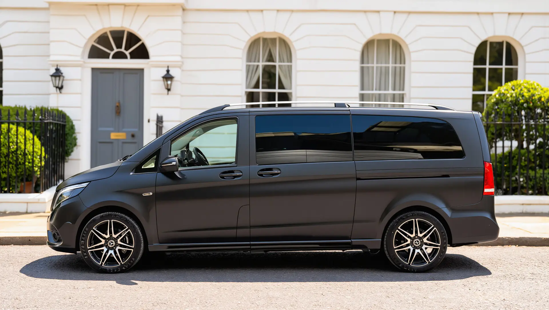 A black Mercedes Senzati V Class van is parked on a residential street in front of a white building with large windows and a grey door.