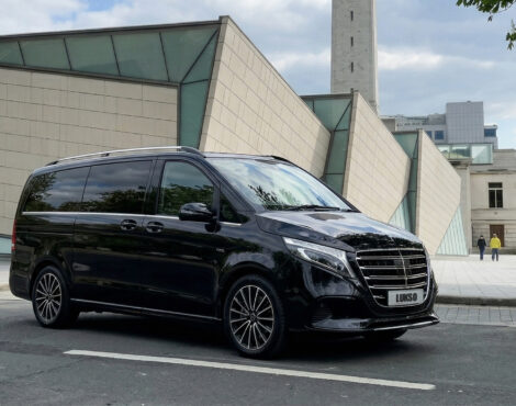 A black Mercedes Senzati V Class van is parked on a city street in front of a modern building with glass and beige panelling.