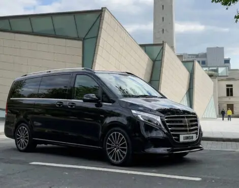 A black Mercedes Senzati V Class van is parked on a city street in front of a modern building with glass and beige panelling.