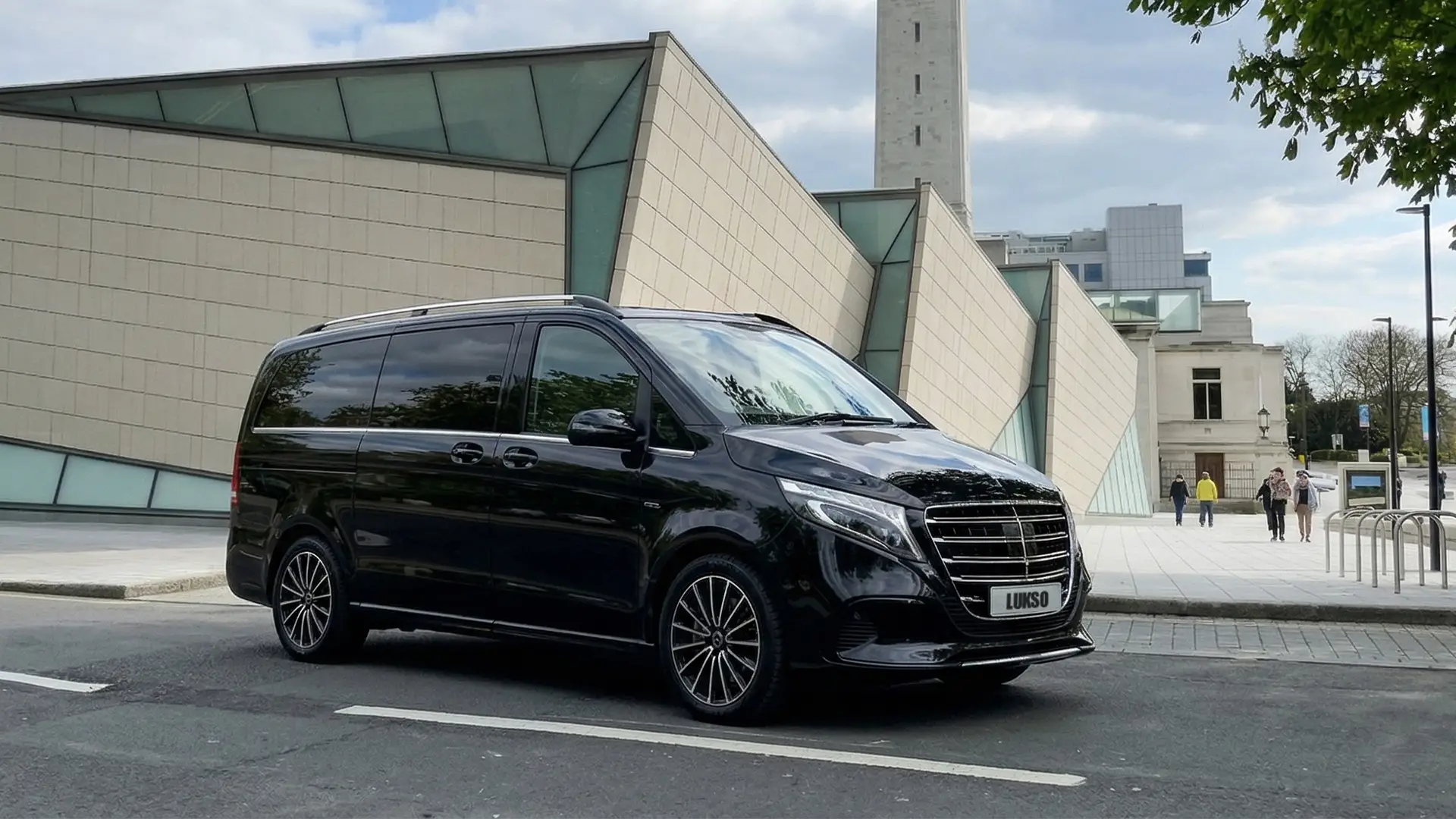 A black Mercedes Senzati V Class van is parked on a city street in front of a modern building with glass and beige panelling.