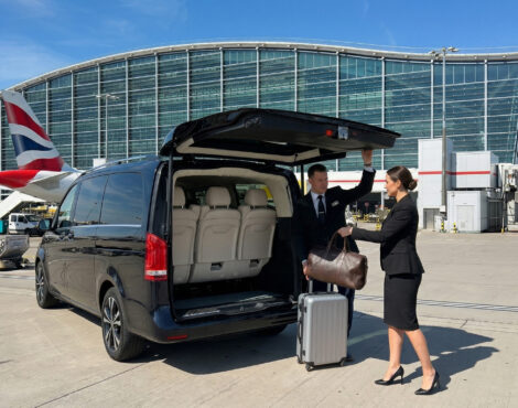 Two people in business attire unload luggage from a Mercedes Senzati V-Class van at an airport, with a terminal building and a British Airways aeroplane visible in the background.