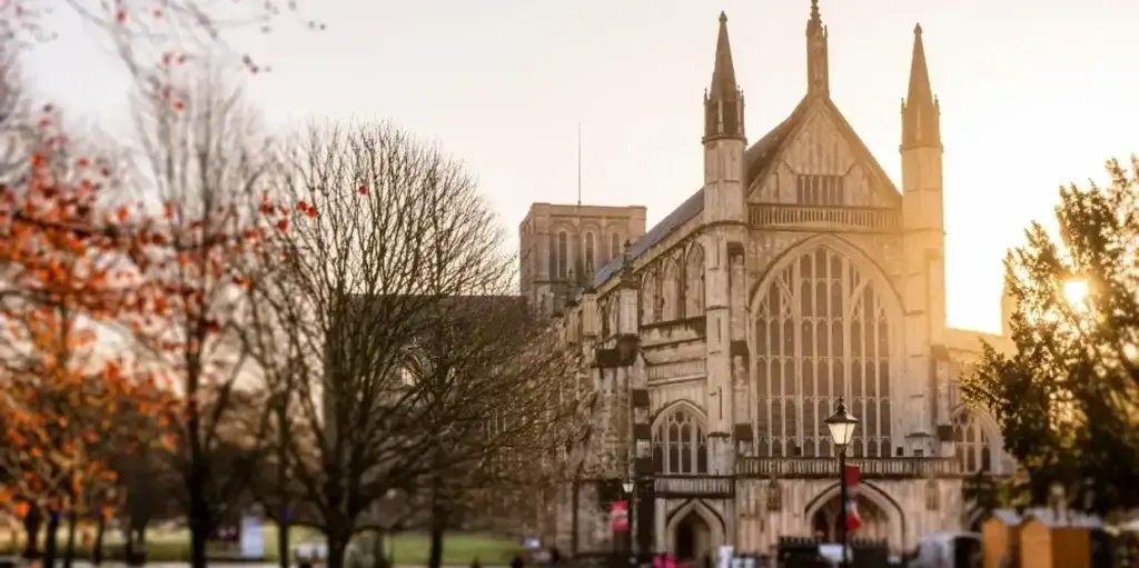A large Gothic-style cathedral with tall spires is illuminated by the setting sun, surrounded by bare trees and a grassy area—an elegant spot to admire while your chauffeur Southampton awaits nearby.