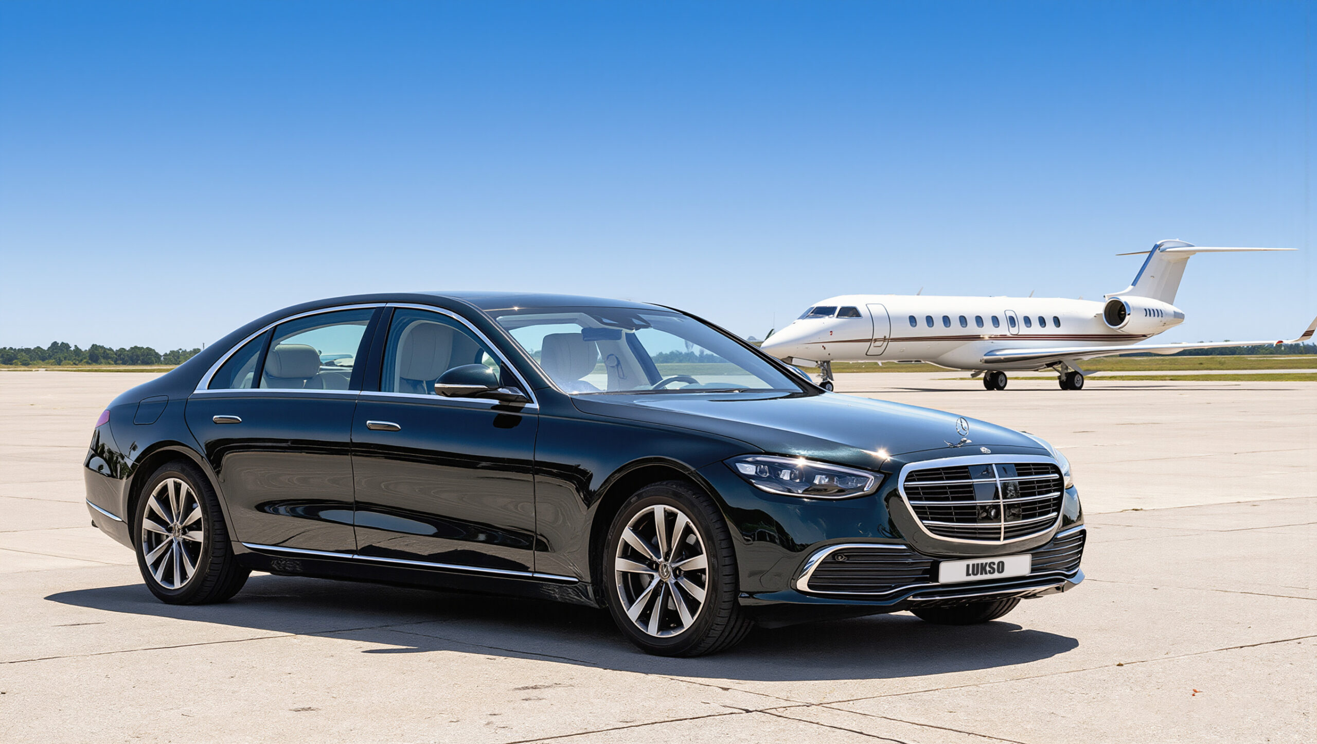 A black Mercedes S-Class (LWB) luxury saloon is parked on an airport apron with a private jet in the background under a clear blue sky.