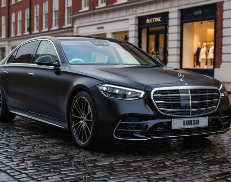 A black Mercedes S Class (LWB) saloon is parked on a wet cobblestone street in front of upmarket shops on a cloudy day.
