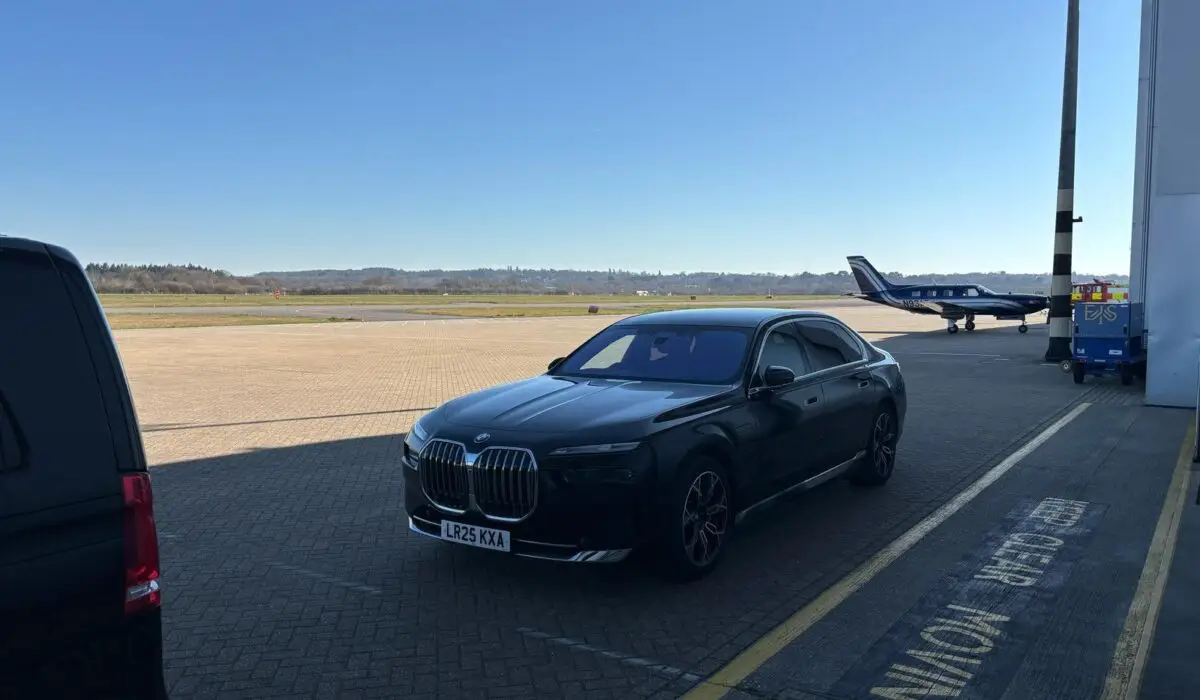 A black BMW saloon, operated by a chauffeur Portsmouth service, is parked on an airport apron near a private jet under a clear blue sky.