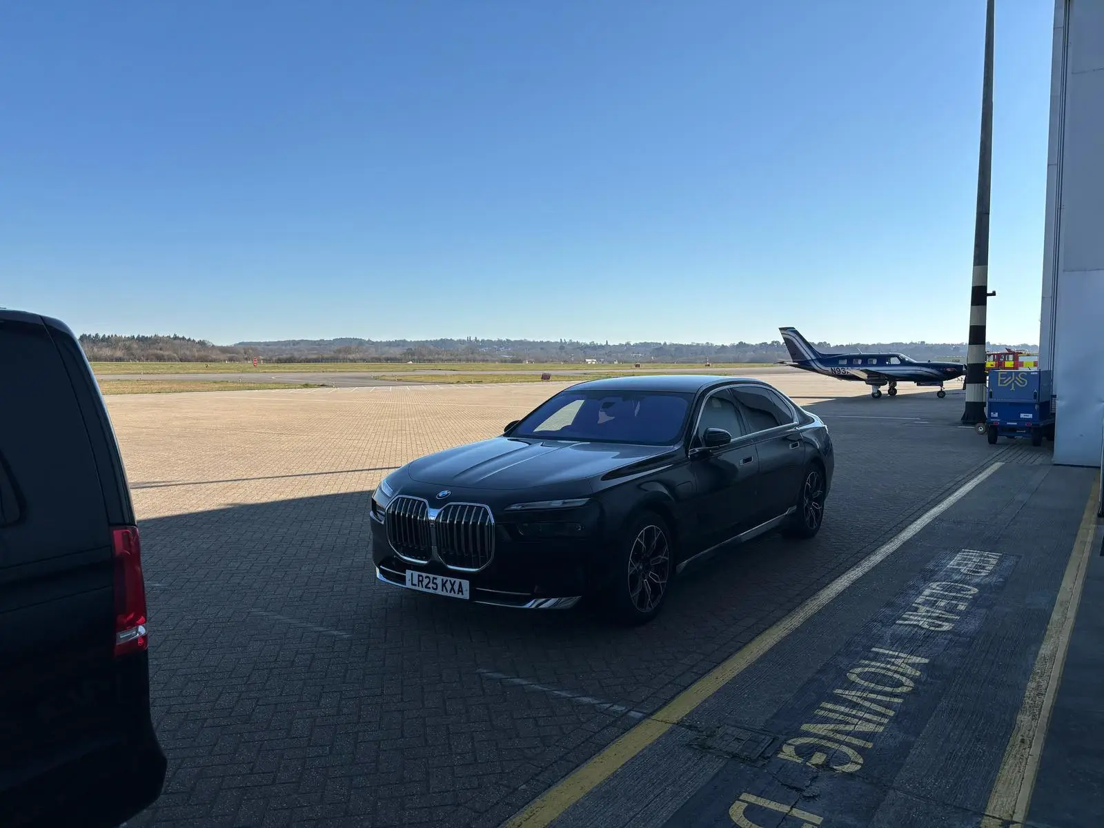 A black BMW saloon, operated by a chauffeur Portsmouth service, is parked on an airport apron near a private jet under a clear blue sky.