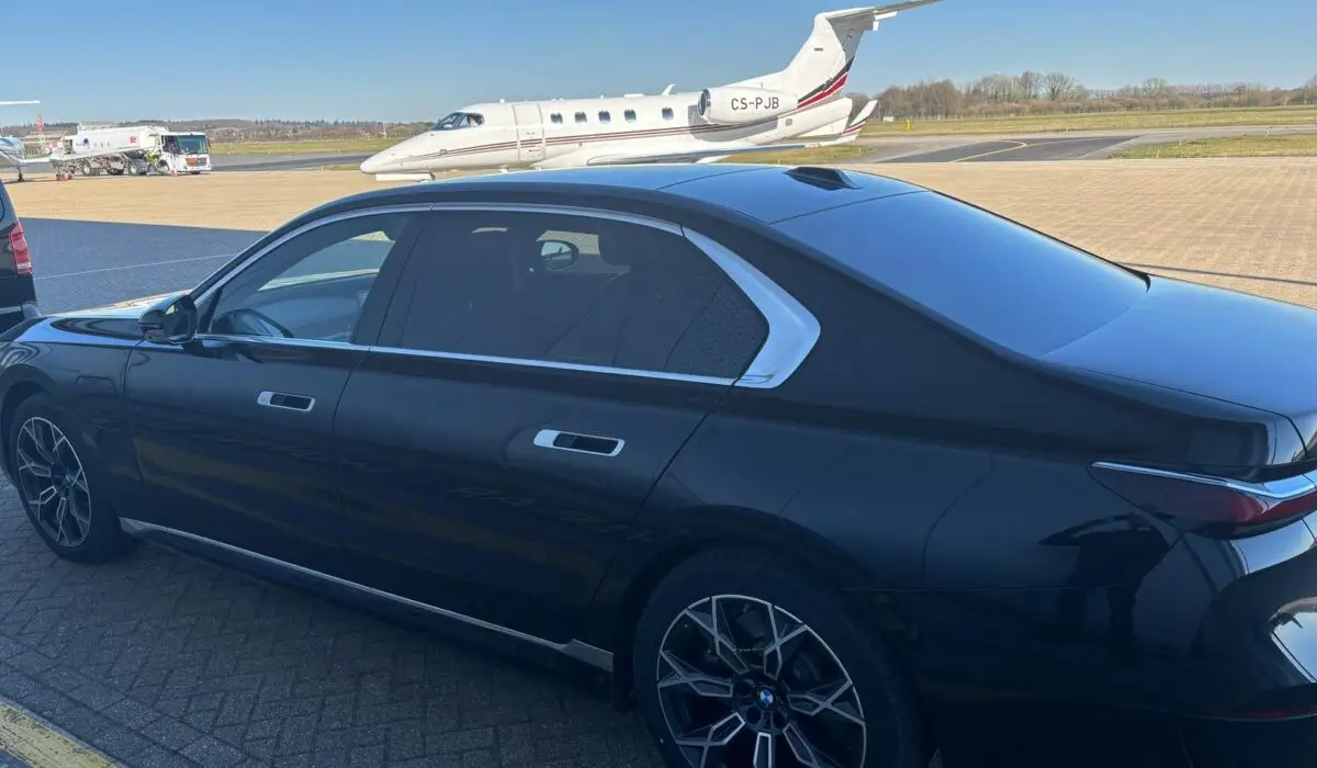 A black luxury saloon, arranged by a chauffeur Portsmouth service, is parked on an airport apron with a white private jet in the background under a clear blue sky.