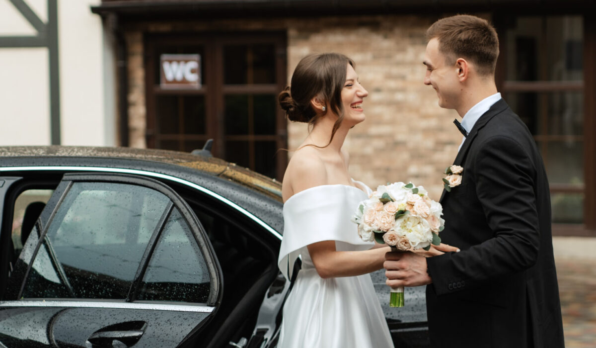 A bride and groom stand smiling at each other beside a car, their wedding chauffeur waiting nearby. The bride holds a bouquet of flowers and wears an off-the-shoulder white dress; the groom wears a black suit with a boutonniere.