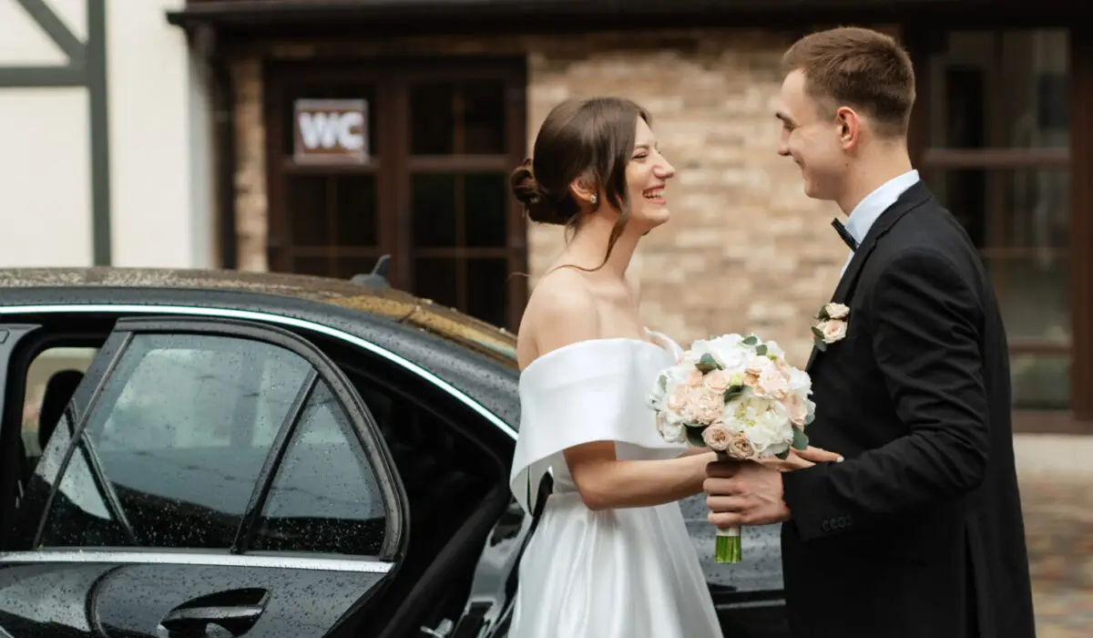 A bride and groom stand smiling at each other beside a car, their wedding chauffeur waiting nearby. The bride holds a bouquet of flowers and wears an off-the-shoulder white dress; the groom wears a black suit with a boutonniere.