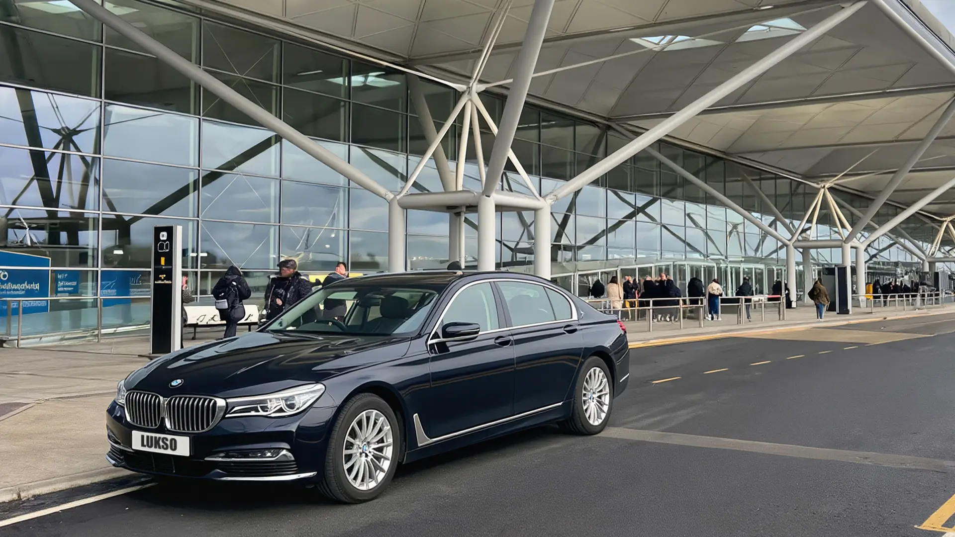 A black BMW saloon with the number plate LUKSO is parked outside Gatwick Airport’s modern terminal, featuring glass windows and a covered entrance.