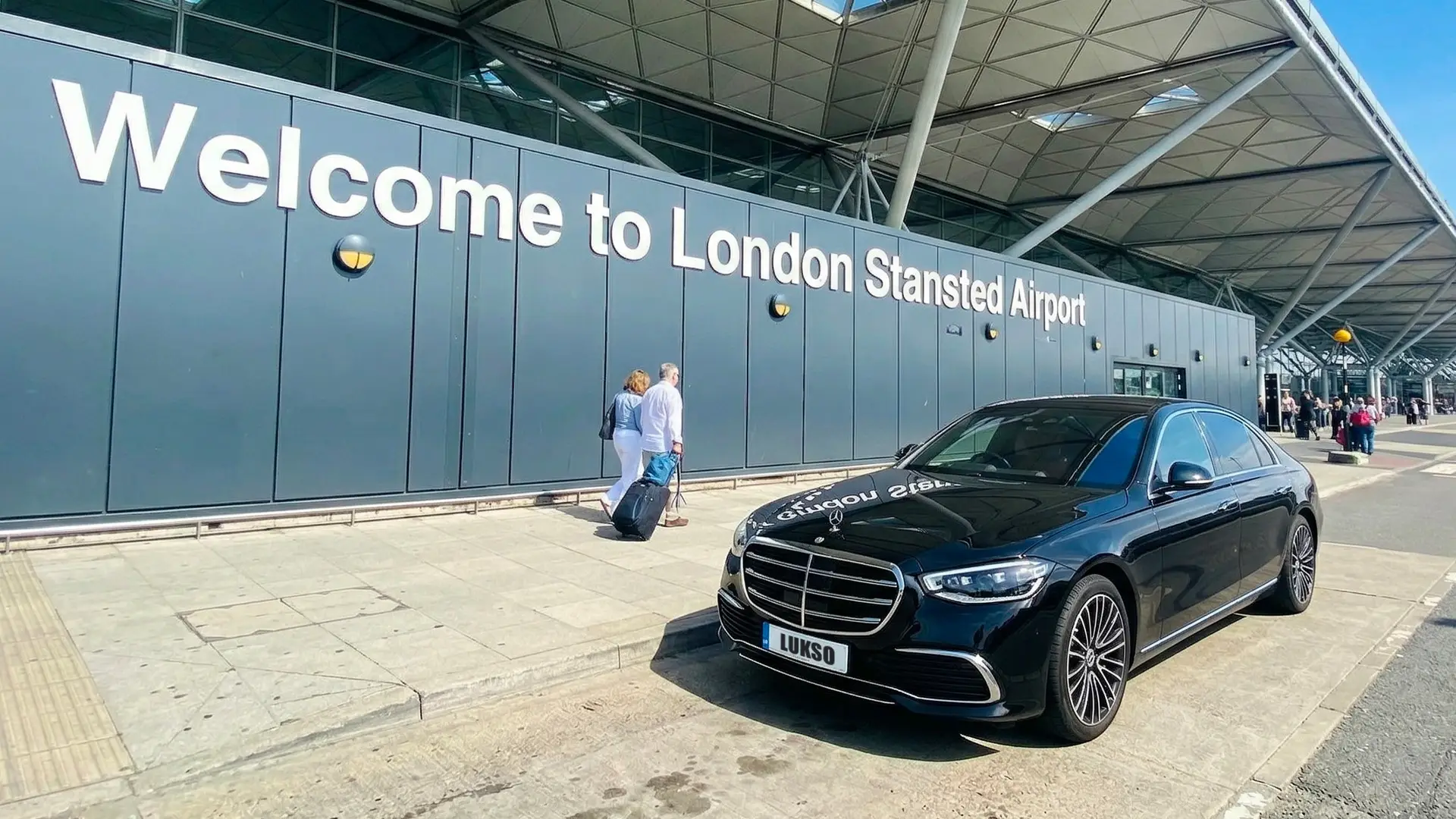 A black Mercedes-Benz saloon is parked in front of the Welcome to Stansted Airport sign, with people walking on the pavement nearby at this busy UK airport.