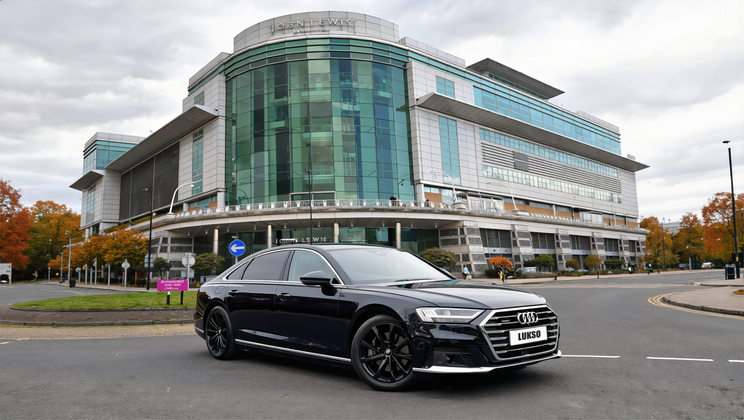 A black Audi A8L saloon is parked on a street in front of a large, modern glass office building under a cloudy sky.